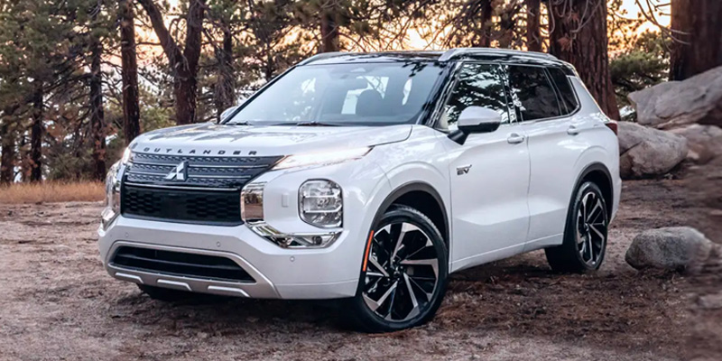 2026 Mitsubishi Outlander SUV in white parked on a forest trail, featuring a bold front grille, modern styling, and tall trees and rocks in a natural outdoor setting.