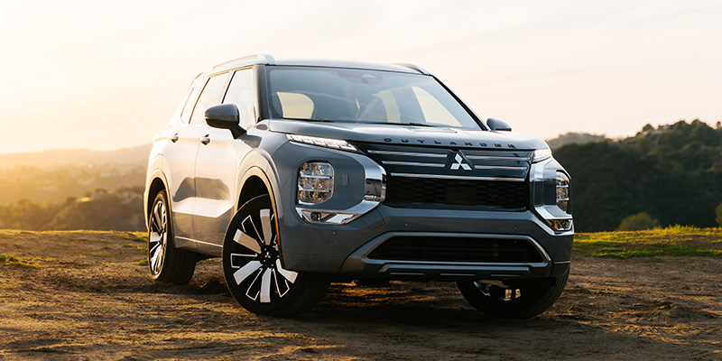New 2026 Mitsubishi SUV parked on a dirt overlook at sunset, featuring a modern gray exterior, bold front grille, and scenic hills in the background.